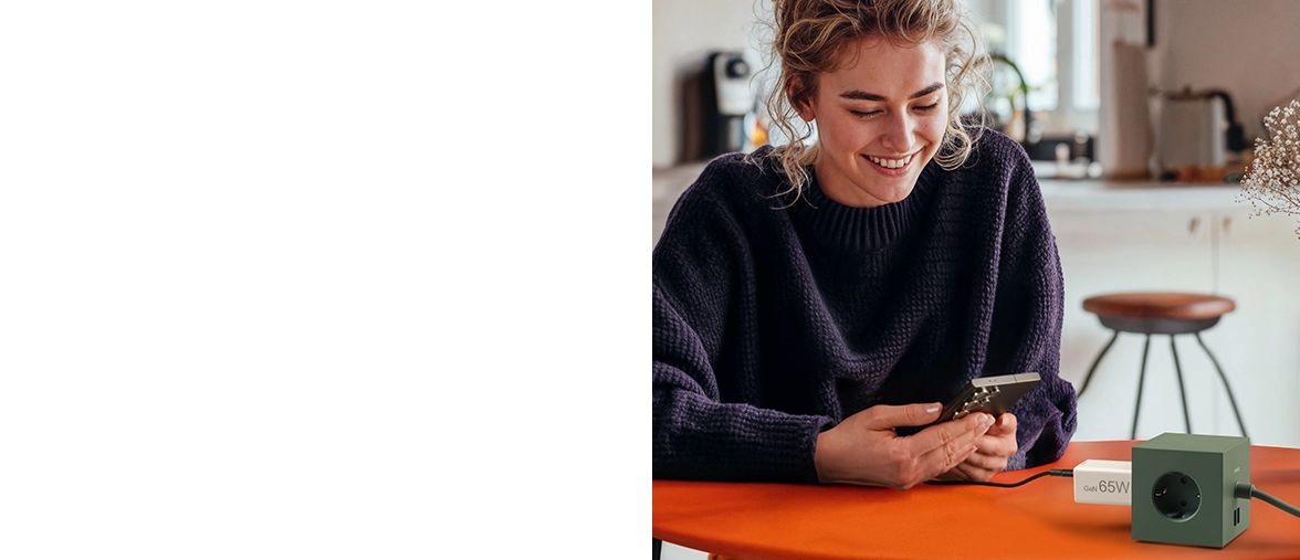 A young woman sits at a table in the kitchen and uses her smartphone while it is being charged via a Hama quick charger.