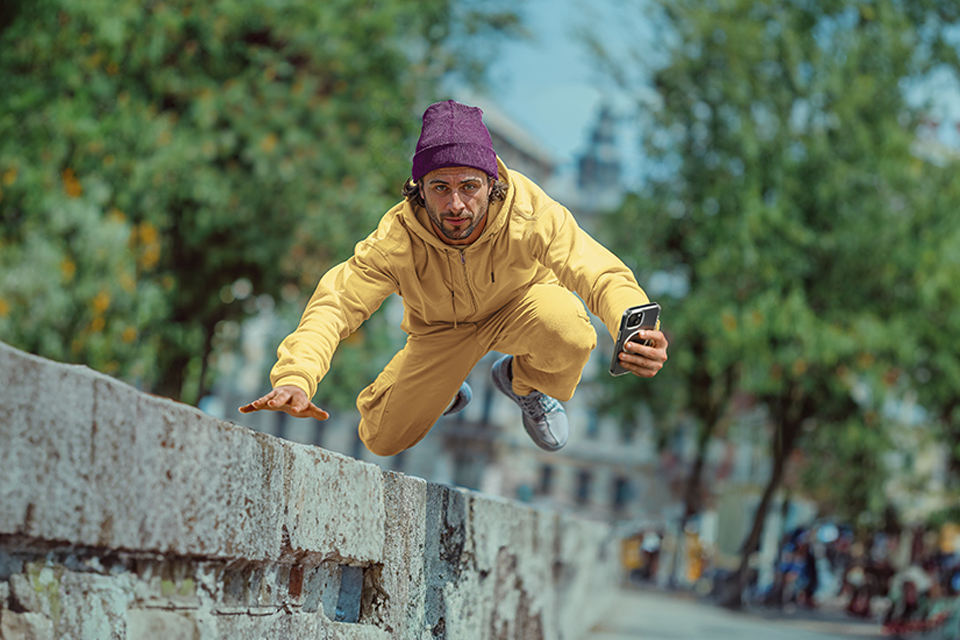 A young man in a yellow jumper and trousers and a purple cap jumps over a wall, holding his smartphone with a Hama cover made of D3O material and looking directly into the camera.