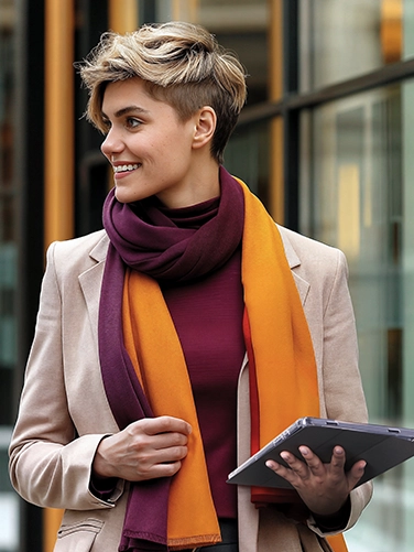 A young woman is out and about in the city, dressed in a beige jacket and an orange scarf. The Apple iPad 10.9’ in her left hand is optimally protected with the Hama “Extreme Protect” tablet sleeve.