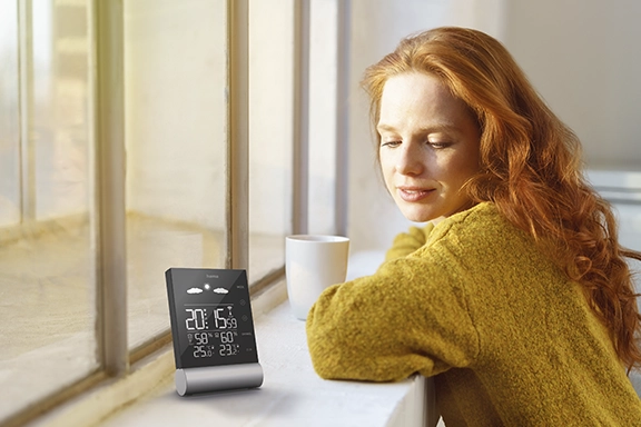 A young woman sits at the windowsill with a cup of tea and looks at the "Black Line" weather station from Hama