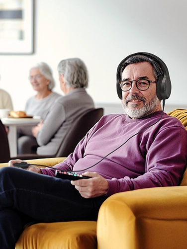 An elderly man sits in a comfortable leather armchair in the foreground wearing Hama TV headphones. In the background, several older women are chatting at a table.