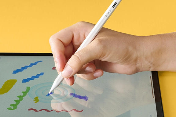 A tablet with an open drawing programme lies on a yellow background. One hand holds the Hama tablet pen ‘Scribble’ and draws different coloured scribbles in the app.