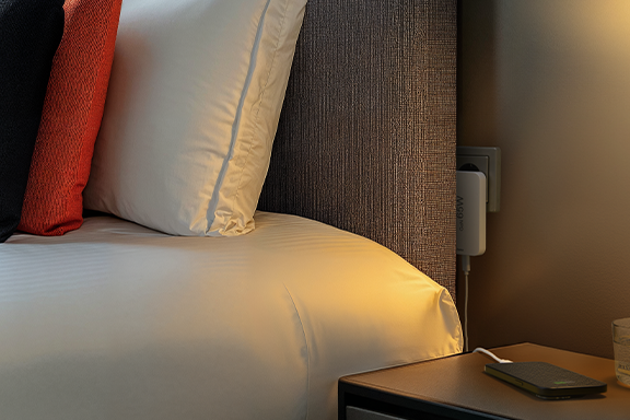 The flat Hama quick charger is plugged into a socket on the wall behind the bed in a bedroom. On a bedside cabinet next to the bed is a smartphone that is charged via the fast charger.
