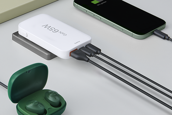 The Hama quick charger with 65 watts, a smartphone, tablet and Bluetooth headphones, which are charged simultaneously via the mini quick charger, are lying on a desk.