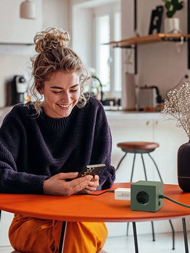 Young woman sitting at a dining table and operating her smartphone while it is being charged with the Hama fast charger, 65W