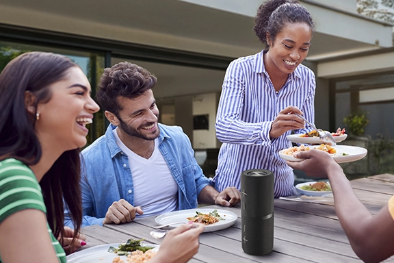 Several people are sitting together at a barbecue in the garden and the Bluetooth Box 4.0 is on the table.