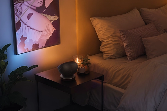 The smart LED table lamp from Hama stands on a side table in the bedroom and indirectly illuminates the wall behind the bed with pleasantly warm light.