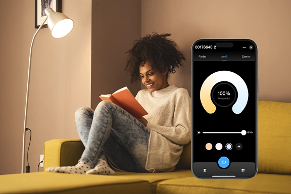 In the background, a young woman sits comfortably on the sofa under a reading lamp with the smart Hama WLAN LED lamp and reads a book. On the right-hand side of the picture you can see a smartphone with the Smart Home app open