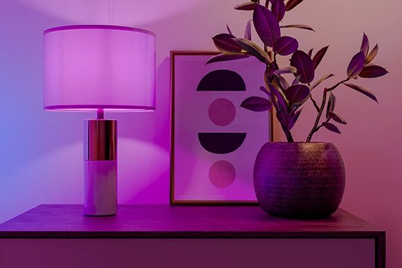 On a wooden sideboard stand a vase, a picture frame and a lamp that sets the smart Hama WLAN lamp in a harmonious purple-coloured light.