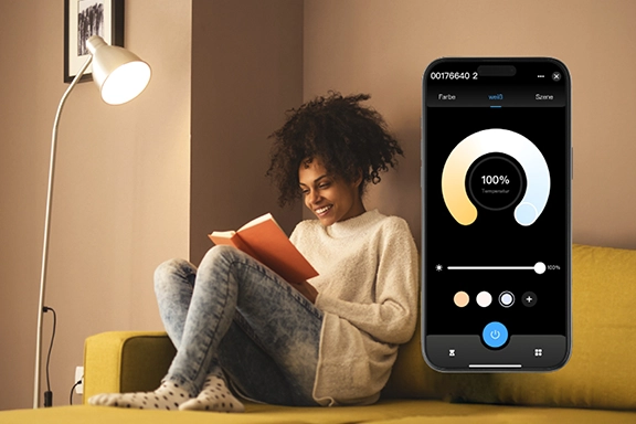 On the right-hand side, you can see a smartphone with the Smart Home app open. On the left and in the background, a woman sits relaxed on the sofa and reads a book under a floor lamp next to her.