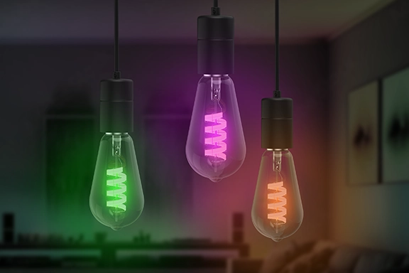 The smart Hama LED lamp hangs three times side by side from the top of the picture in the foreground. Each bulb lights up differently: green, pink and orange. A darkened and blurred living room can be seen in the background.