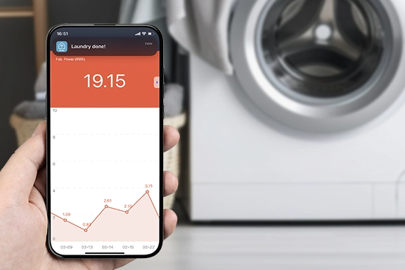In the foreground, a hand holds a smartphone on which the smart home app is open and shows temperature statistics. A laundry room with a washing machine can be seen in the background.