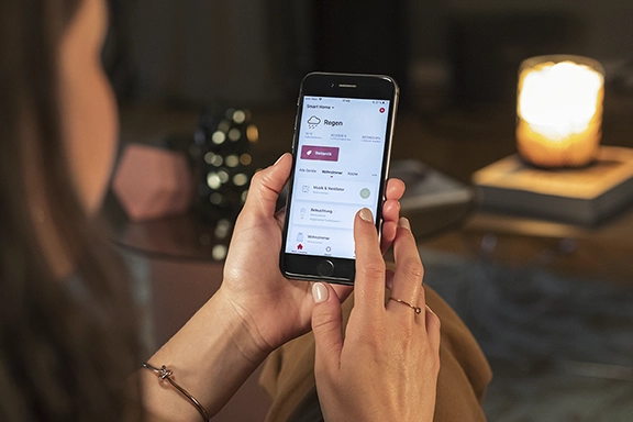 A young woman sits in a dark living room and controls the temperature in the room using the Hama Smart Home app.