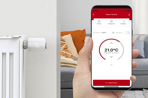 In the foreground is a smartphone with the Smart Home app open, which can be used to control the temperature in the living room using a smart radiator thermostat. In the background is a living room with a radiator and installed radiator thermostat.