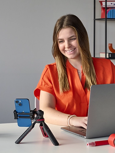 A young woman works on her laptop while streaming on her mobile phone, which is attached to the Hama "Solid" tripod